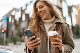 Young woman with curly blonde hair using the phone with a cup of coffee in hands on the city streets, portrait