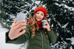 Winter selfie. Beautiful girl in winter clothes takes selfie on smartphone. Woman with coffee takes selfie in winter snowy day