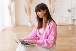 Donna con camicia rosa che scrive sul laptop a casa
