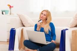 Donna bionda seduta davanti al divano con laptop, con una carta di credito in mano