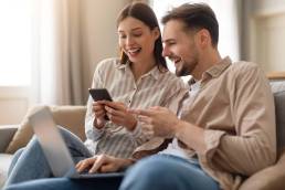 Coppia sorridente che condivide uno smartphone mentre l’uomo ha un laptop sulle ginocchia