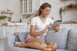 Donna ripresa a figura intera mentre è seduta su un divano con in mano un tablet