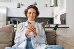 portrait-smiling-woman-chats-smartphone-app-sits-home-sofa-uses-mobile-phone-applica