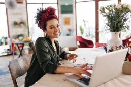 Donna giovane con capelli ricci scuri seduta a un tavolo in un laboratorio luminoso e moderno, mentre disegna al computer e sorride verso la fotocamera