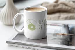 Tazza bianca con logo verde &ldquo;Il Rifugio di Anna&rdquo; su rivista, accanto a vaso bianco
