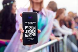 Persona mostra uno smartphone con un QR code per la partecipazione a un evento, sullo sfondo folla sfocata a un concerto o festival
