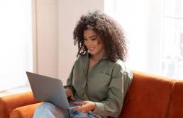 ragazza con una folta chioma riccia, sorridente mentre lavora al computer seduta sul suo divano color terracotta