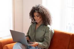 ragazza con una folta chioma riccia, sorridente mentre lavora al computer seduta sul suo divano color terracotta