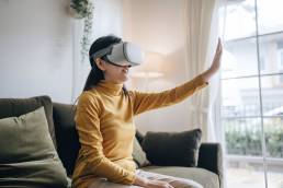 ragazza seduta sul divano di casa mentre indossa un visore per la realt&agrave; virtuale