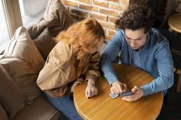 Un ragazzo e una ragazza seduti a un tavolo di legno scansionano un codice QR o scattano una foto con lo smartphone, in un ambiente accogliente con mattoni a vista.