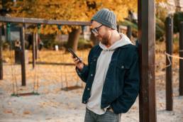 Standing with smartphone. Beautiful man in warm clothes is outdoors in the park