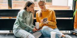 Due donne sedute su una panca in un bar guardano insieme lo smartphone e sorridono, con bicchieri da caffè davanti a loro.