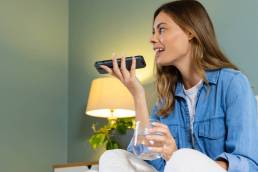 Donna che si rilassa a casa, utilizzando l'assistente vocale dello smartphone, con un bicchiere d'acqua in mano.