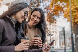 Two cheerful young girls using smartphone while sitting at the bench outdoors