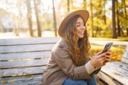 Young smiling woman sitting on bench in autumn park using phone in stylish clothes happy mood