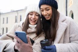 Female friends looking at smartphone and holding coffee cups outdoors after reuniting