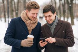 Two men in winter coats looking at phone with coffee outdoors